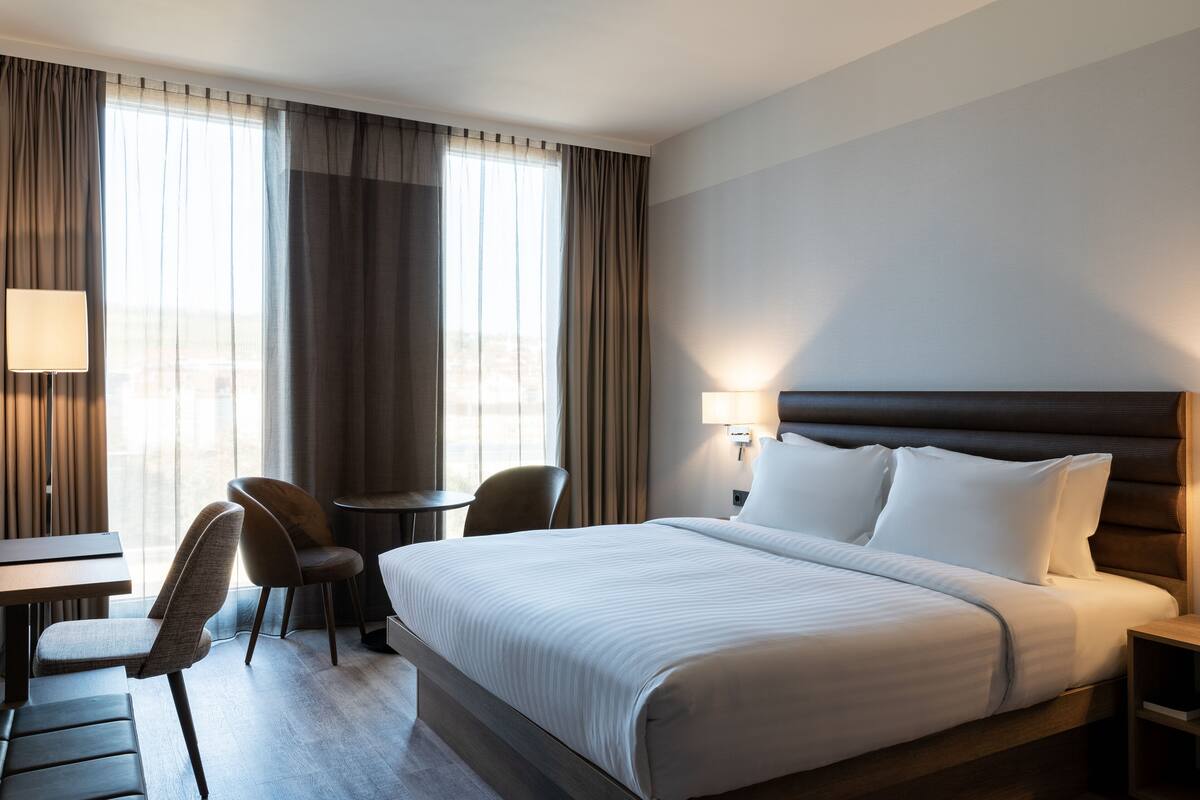 Une chambre d’hôtel avec un grand lit, un bureau, deux chaises, une lampe et une vue sur la ville par la fenêtre.