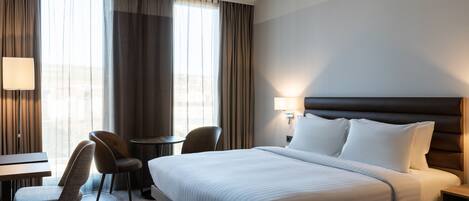 Habitación de hotel con una cama grande, un escritorio, dos sillas, una lámpara y vista a la ciudad por la ventana.
