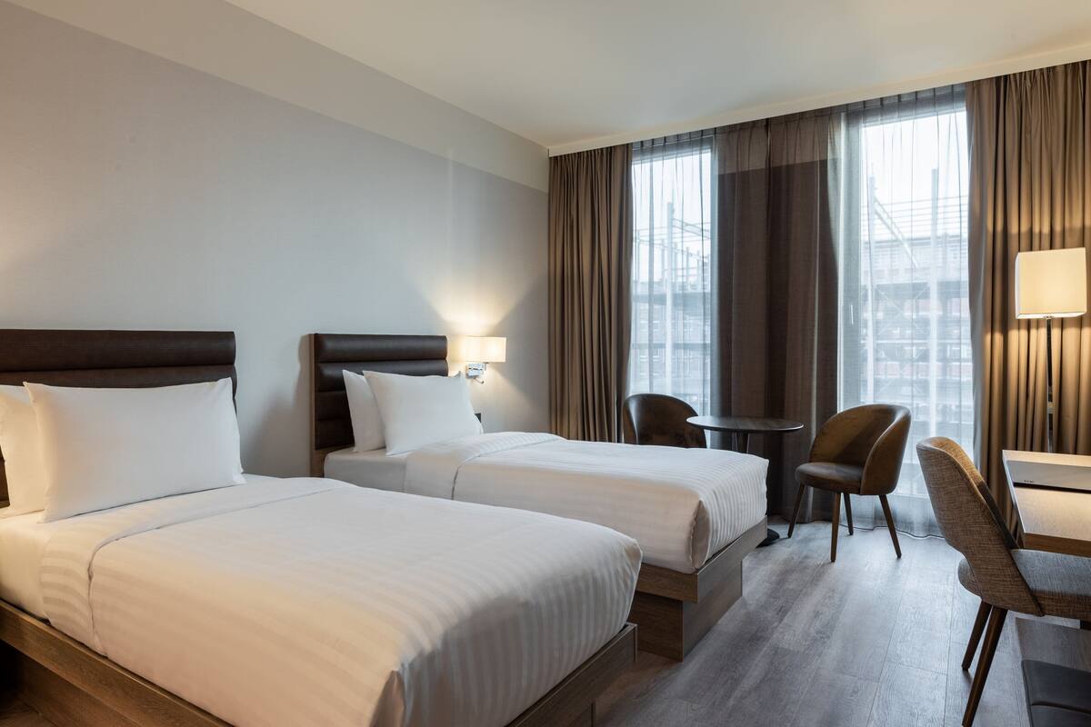 Une chambre d’hôtel avec deux lits, un bureau, une chaise et une fenêtre avec des rideaux.