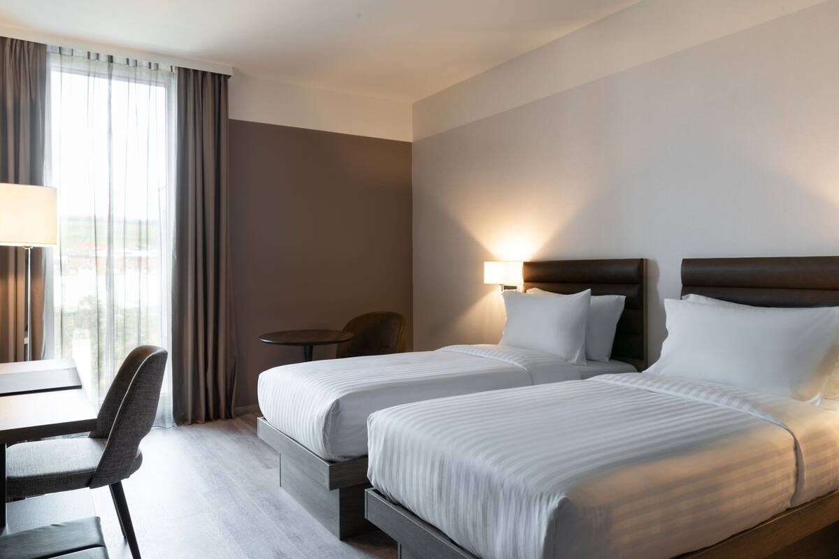 A hotel room with two beds, a desk, a chair, and a lamp.