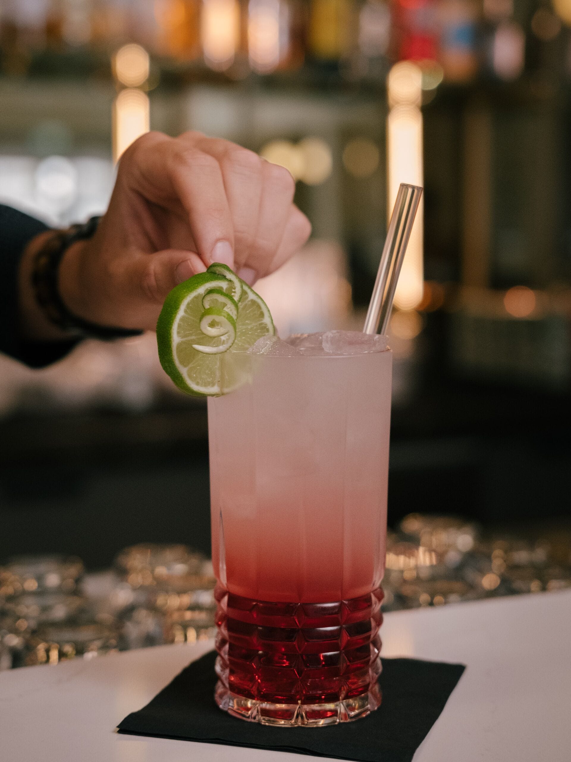 A hand holding a lime slice above a tall glass with a gradient of red to pink liquid, likely a cocktail, with a metal straw.