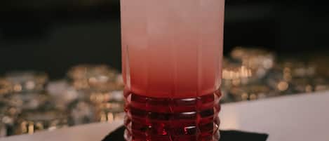 Una mano sosteniendo un gajo de lima sobre un vaso alto con un degradado de color rojo a rosa, probablemente un cóctel, con una pajita de metal.