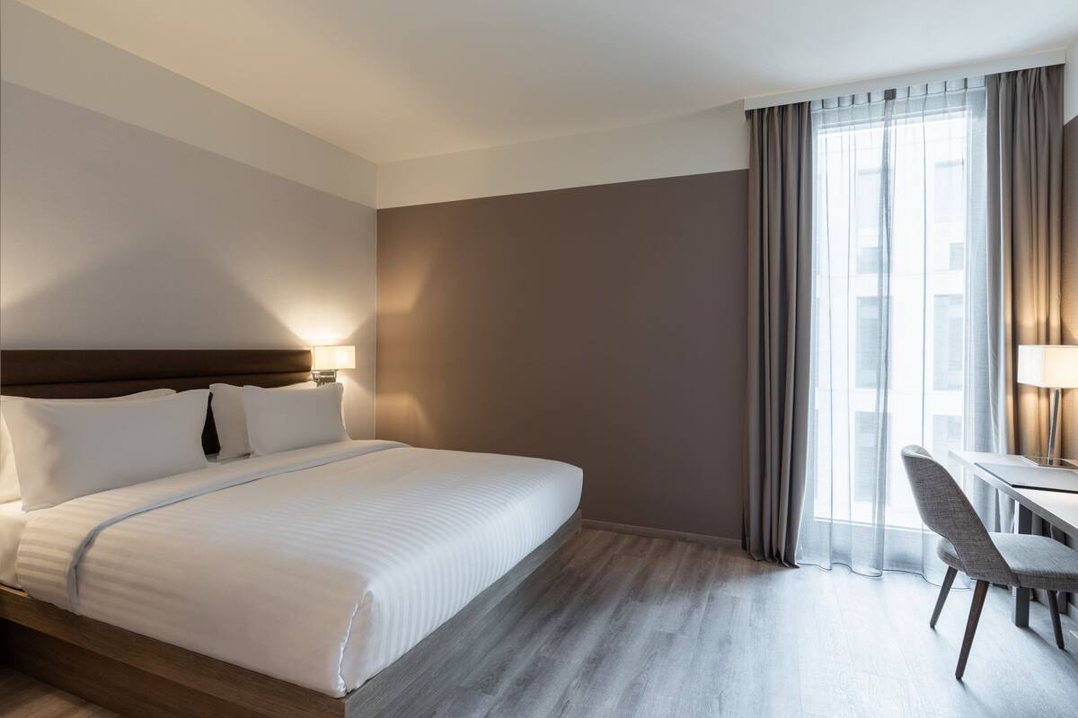 A hotel room with a large bed, a desk with a chair, and a window with curtains.