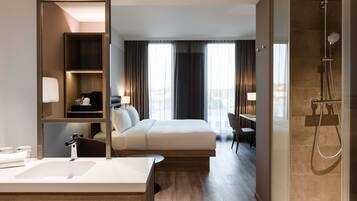 Habitación de hotel con cama, escritorio, ducha, baño con lavamanos y vistas a la ciudad a través de amplias ventanas.