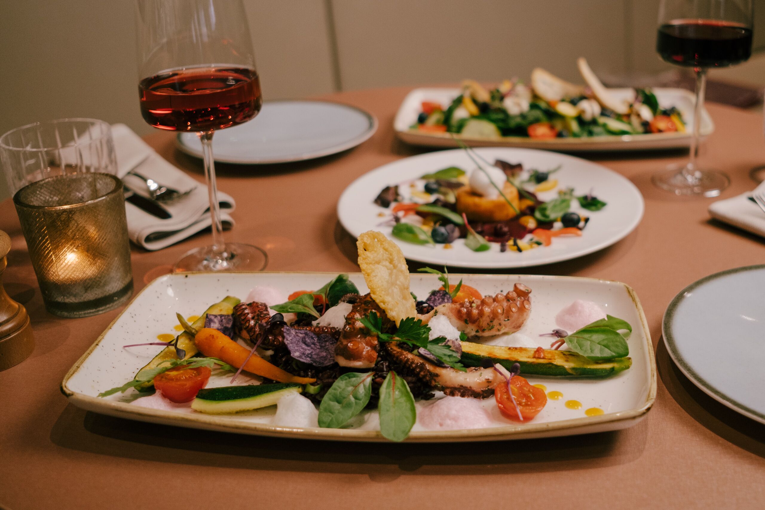 A dining table with plates of food, glasses of red wine, and a candle.