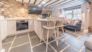 A kitchen with a stone wall, wooden beams, and a stone floor. There is a dining area with a sofa and a TV. - Old Toffee Shop (Ambleside)