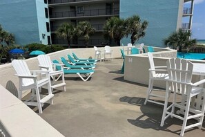 A poolside area with white and turquoise lounge chairs, a small table, and a view of the ocean. - 🏖️ Affordable Cozy Beachfront | FREE Beach Chairs | Early 3PM Check-In (Panama City Beach)