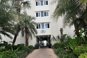 A building entrance with a sign, surrounded by palm trees and greenery. - 🏖️ Affordable Cozy Beachfront | FREE Beach Chairs | Early 3PM Check-In (Panama City Beach)