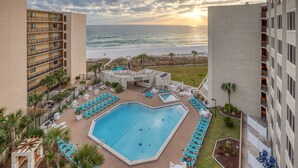 A hotel pool area with a view of the ocean and beach, surrounded by palm trees and lounge chairs.