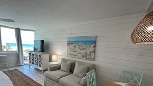 A living room with a sofa, a television, a painting of a beach scene, a table with woven placemats, and a view of the ocean through large windows.