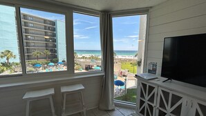 A balcony with a TV, two white bar stools, and a view of a beach and hotel buildings.