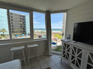 A balcony with a TV, two white bar stools, and a view of a beach and hotel buildings.