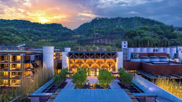 A modern building with a geometric facade, surrounded by greenery and a hilly landscape.