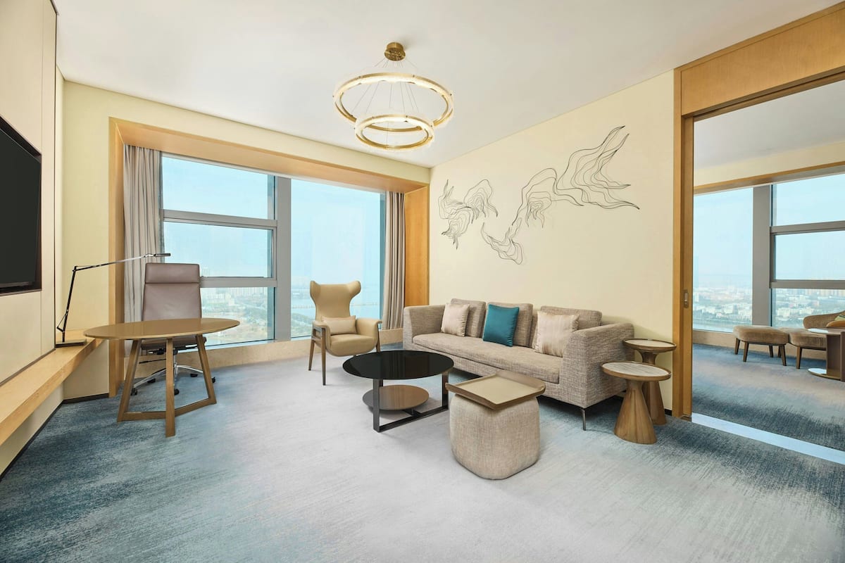 A modern hotel room with a sofa, armchairs, a coffee table, a TV, and large windows offering a city view.