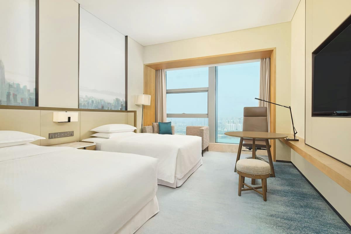 A hotel room with two beds, a desk, a chair, a TV, and a large window with a city view.