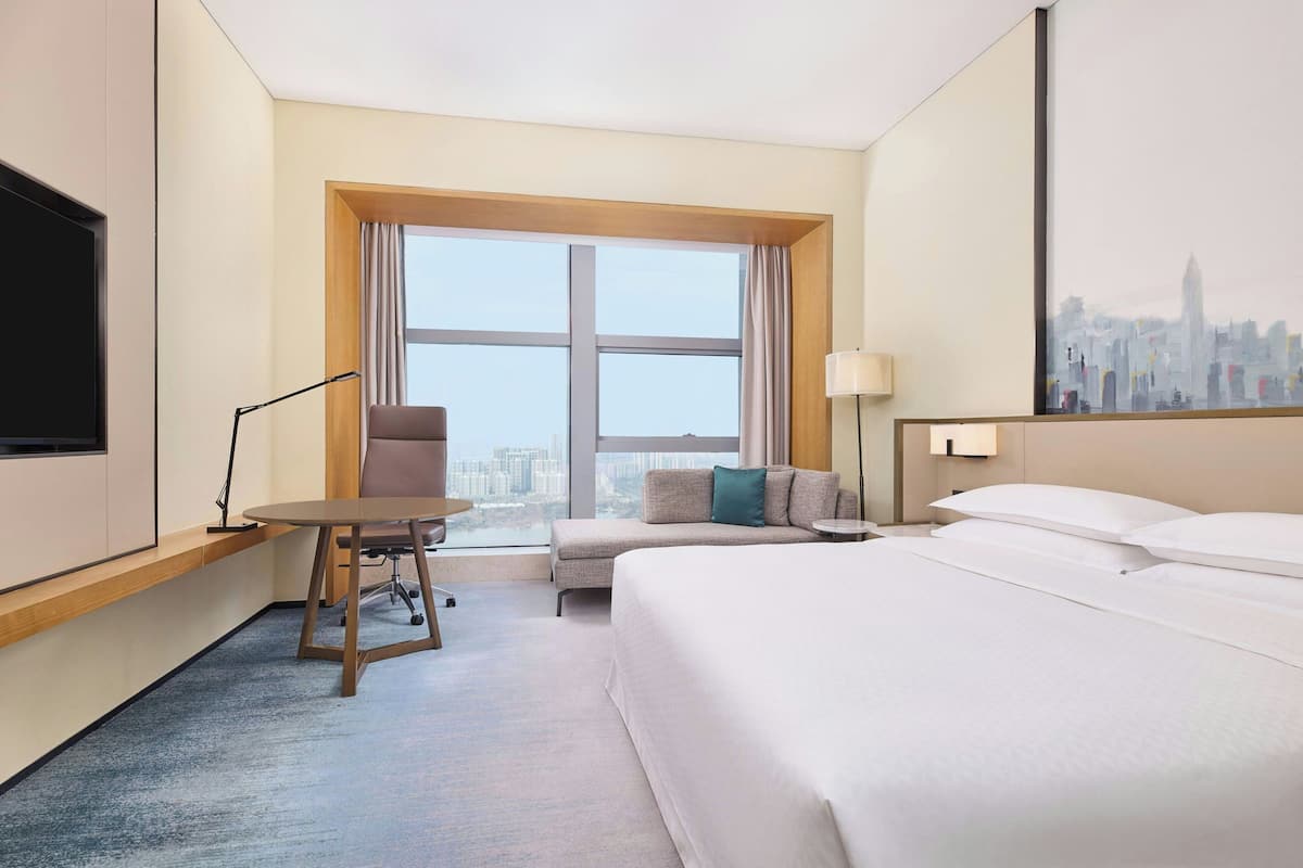 A hotel room with a bed, a desk, a chair, a window with a city view, and a TV.