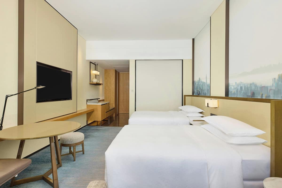 A hotel room with a bed, bedside table, lamp, TV, and a cityscape painting on the wall.