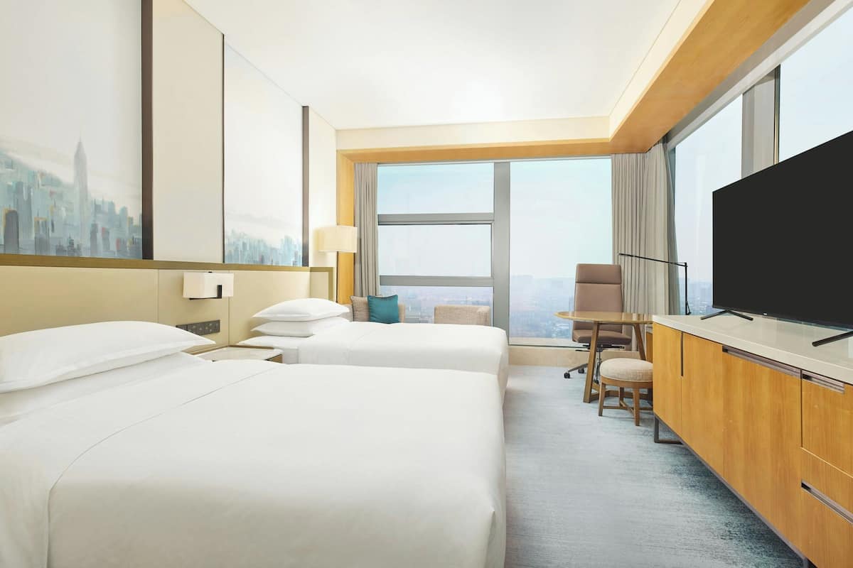 A hotel room with two beds, a TV, a desk, and a large window with a city view.
