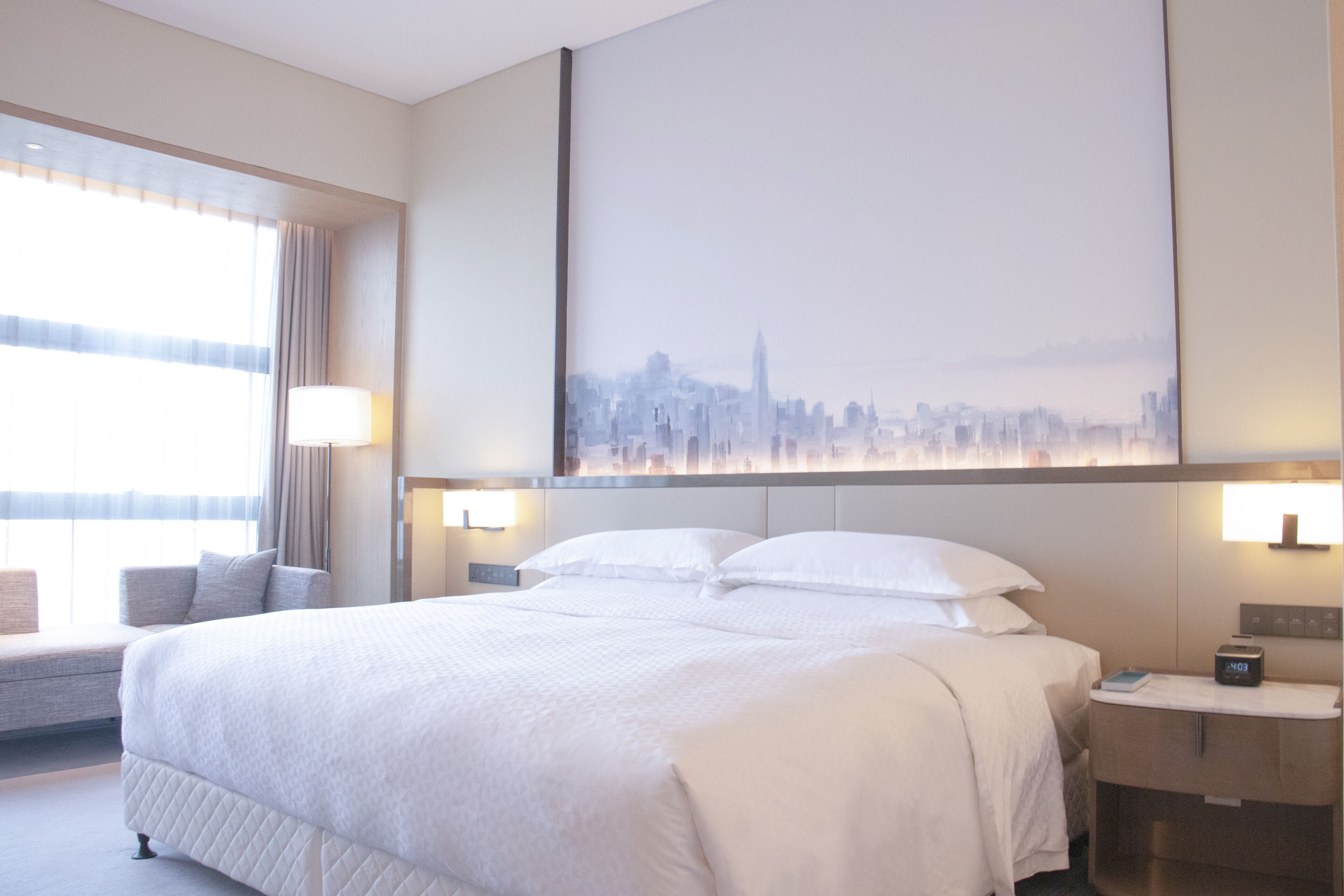 A hotel room with a large bed, a cityscape view, a bedside table with a lamp, and a window with curtains.