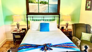 Une chambre d’hôtel avec un lit, deux tables de chevet, une chaise, un tableau représentant une plage et une fenêtre donnant sur des palmiers.