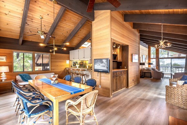 A spacious room with a wooden ceiling, a dining area with a long table and wicker chairs, a kitchen area with cabinets, and a large window providing a view of the outdoors.