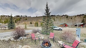 A rustic outdoor seating area with a fire pit, surrounded by rocks and trees, with a car and a small cabin in the background. - Mountain Retreat | Fire Pit | 3 Blocks off Main St. (Grand Lake)