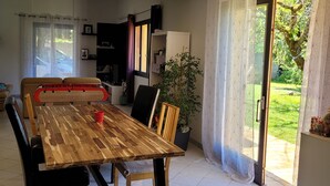 Un coin repas avec une table en bois, des chaises et une tasse rouge. On y trouve également une plante en pot et une porte-fenêtre coulissante donnant sur un jardin.