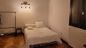 Une chambre à coucher avec un lit, un ventilateur sur pied, un radiateur et une fenêtre dotée d’un cadre foncé.