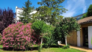 Un jardin avec une pelouse, un arbuste à fleurs roses et un bâtiment doté d’un toit en tuiles.