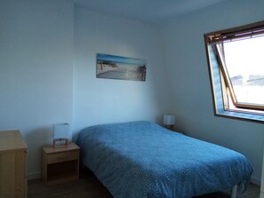 A bedroom with a single bed, a nightstand, a lamp, a small wooden cabinet, and a window with a view of the beach. - Maison Lumineuse 2* Concarneau - 4-minute drive to the beach (Concarneau)