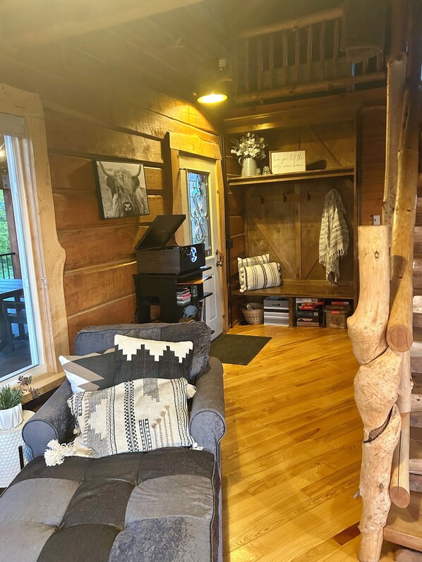 A cozy cabin interior with a wooden floor, a sofa with patterned cushions, a piano, and a rustic staircase.