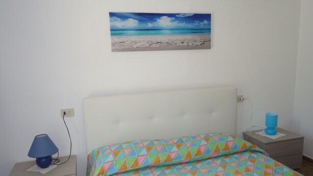 A bedroom with a bed, a nightstand, a lamp, and a wall-mounted picture of a beach. - Holidays at Hydrangea Villa (San Teodoro)