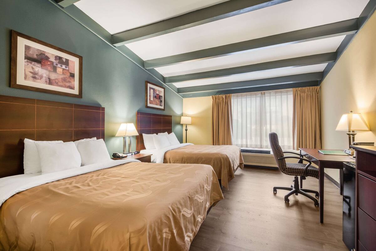 A hotel room with two beds, a desk, and a chair. There are two lamps on the desk and a window with curtains.
