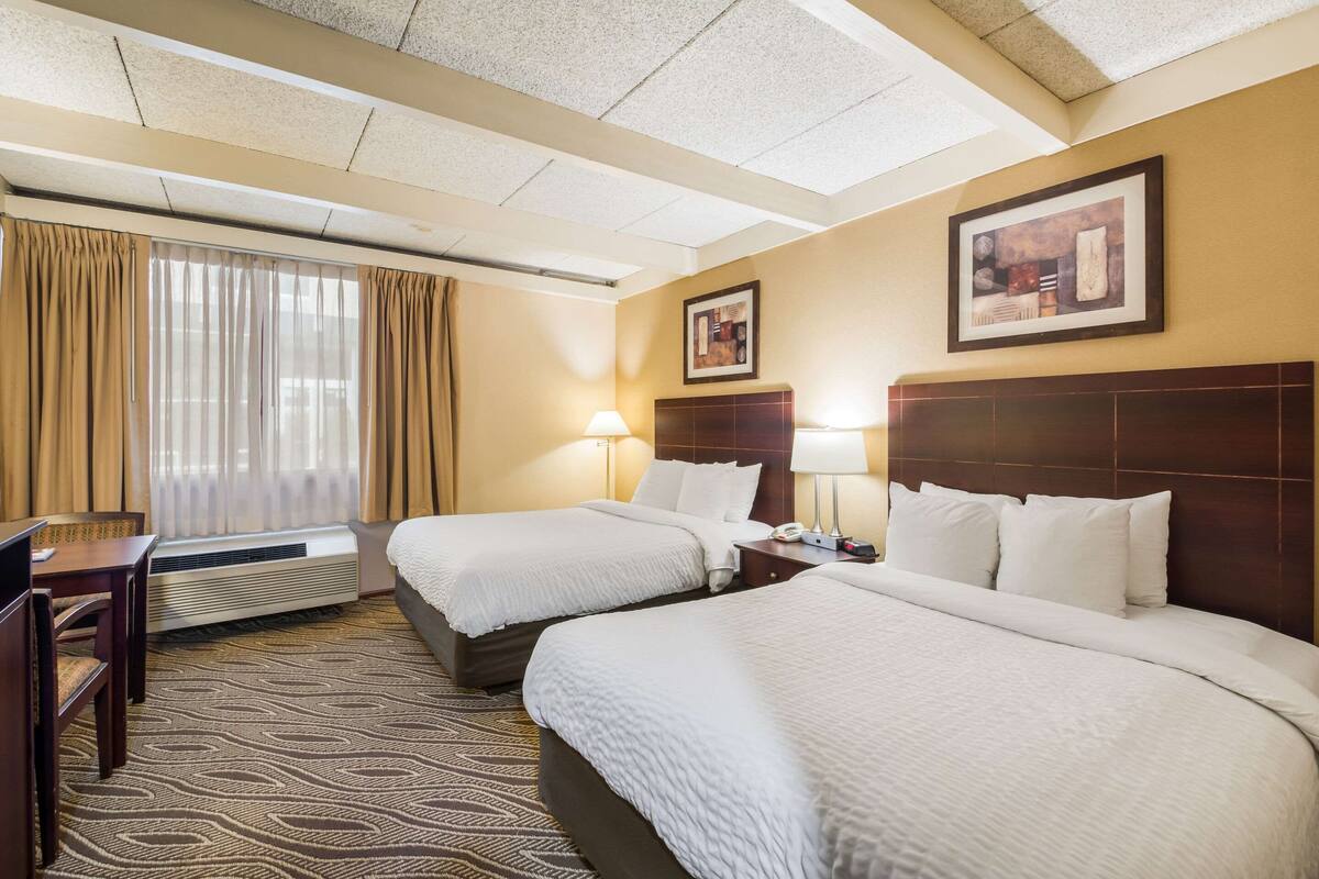 A hotel room with two beds, a desk, and a chair. There is a window with curtains, a wall-mounted lamp, and a framed picture.