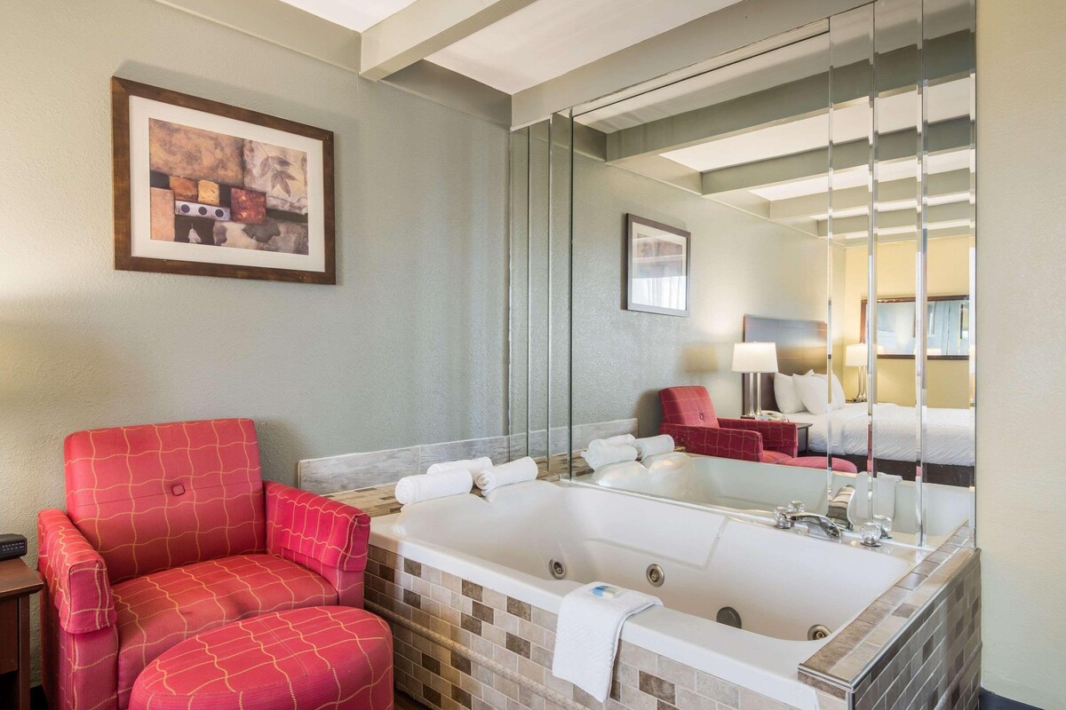 A hotel bathroom with a large bathtub, a red chair, a mirror, and a framed picture on the wall.