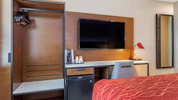 A hotel room with a bed, a desk with a flat-screen TV, a mini-fridge, a coffee maker, and a mirror.