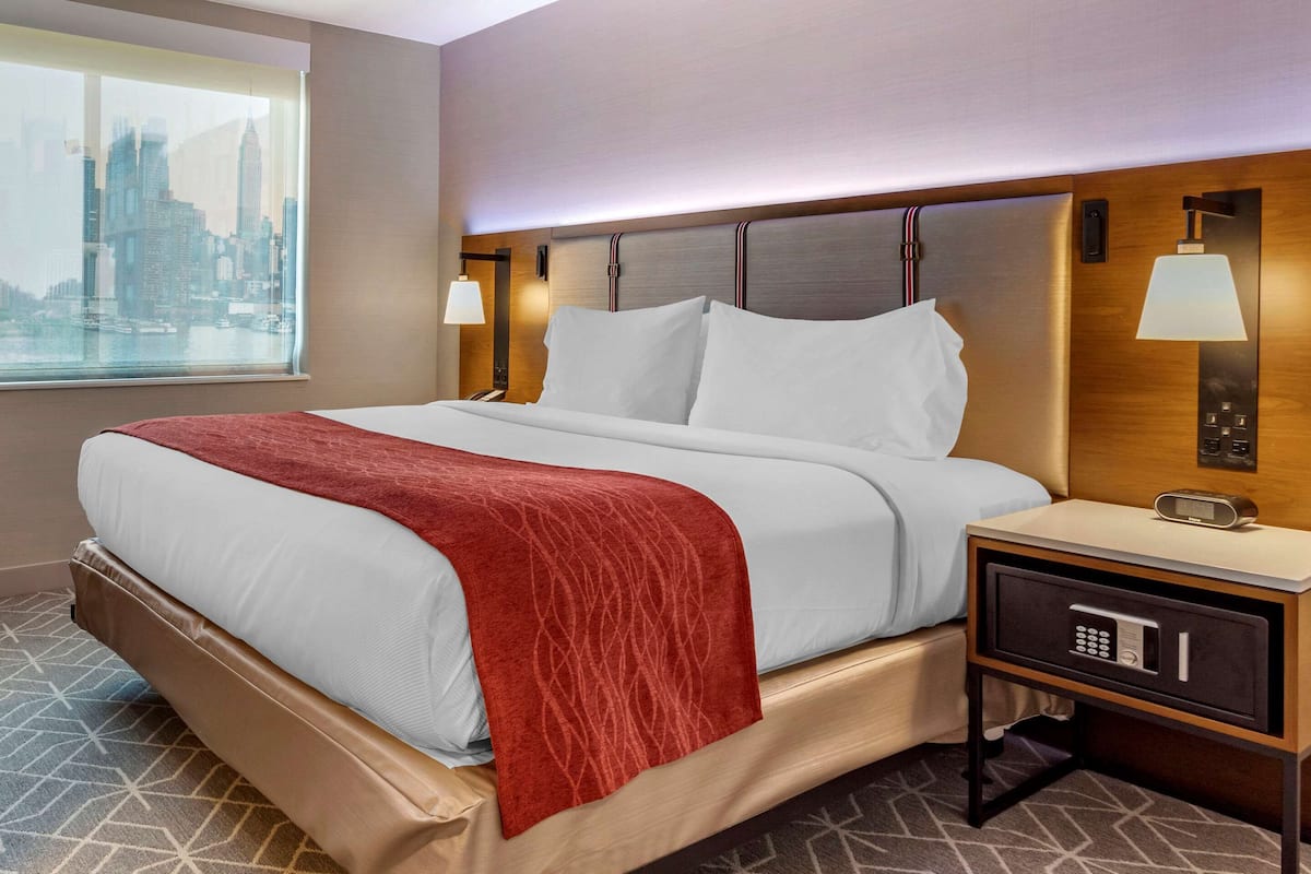 A hotel room with a bed, a nightstand with a lamp, a safe, and a window with a city view.