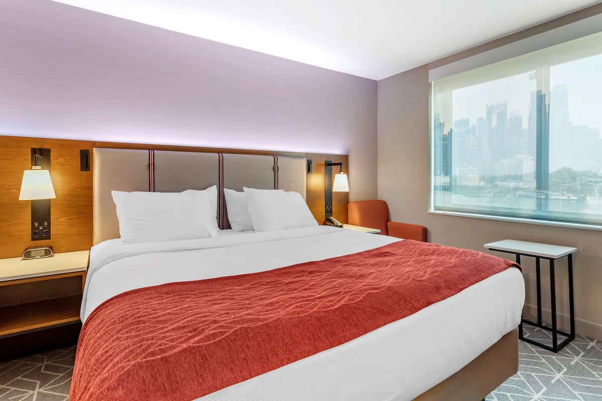 A hotel room with a large bed, two bedside lamps, a nightstand, a small table, and a window with a city view.
