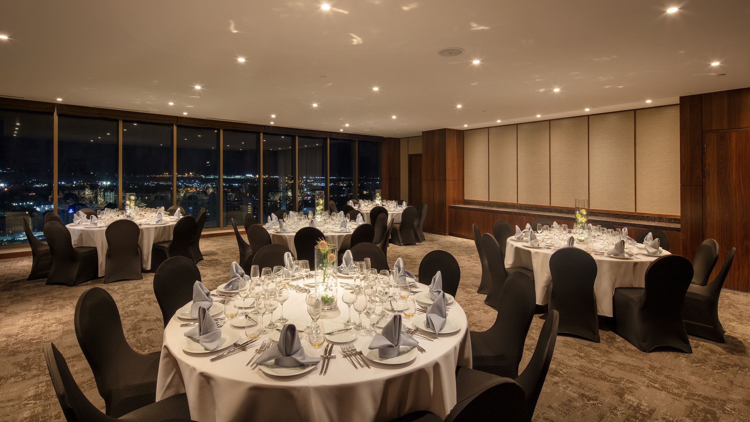 Un salón de conferencias con mesas redondas preparadas para un evento formal, grandes ventanales con vistas a la ciudad y un techo alto con iluminación empotrada.