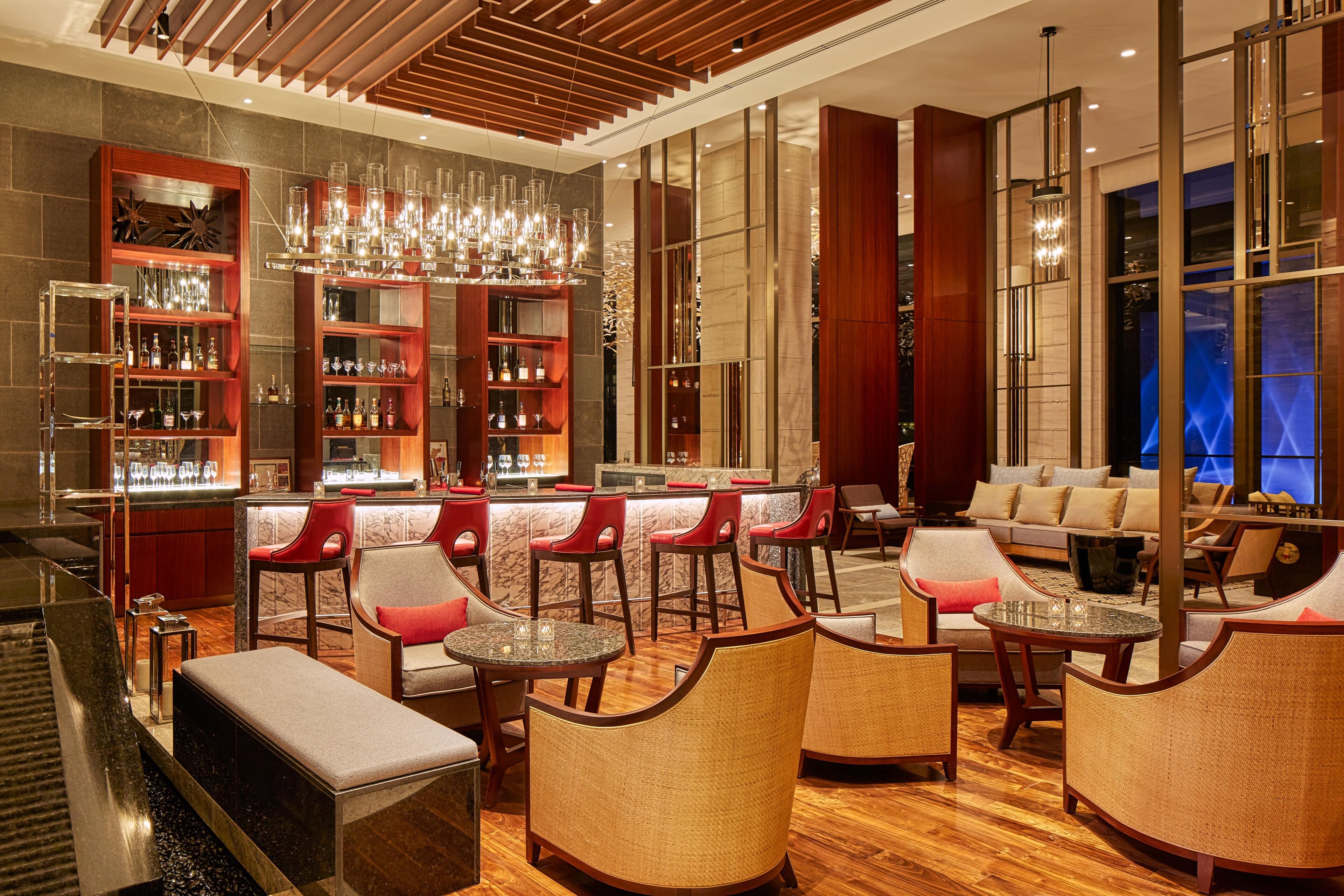 Een moderne bar met houten schappen, rode stoelen, en een zithoek met banken en ronde tafels.