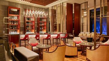 Een moderne bar met houten schappen, rode stoelen, en een zithoek met banken en ronde tafels.