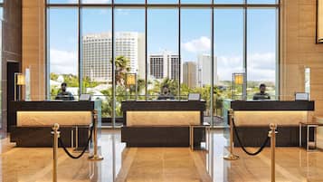 A hotel reception area with three reception desks, each with a computer and a receptionist. The reception area has a modern design with marble flooring and large windows offering a view of the cityscape.