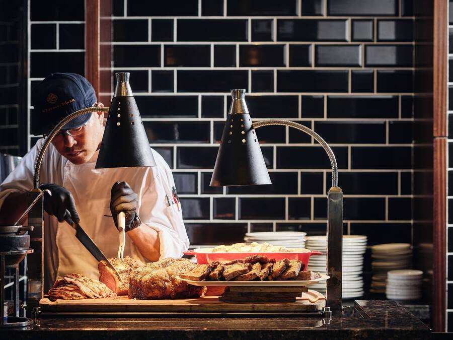 A person slicing meat on a cutting board with a knife, a hookah, and a lamp in a kitchen setting.