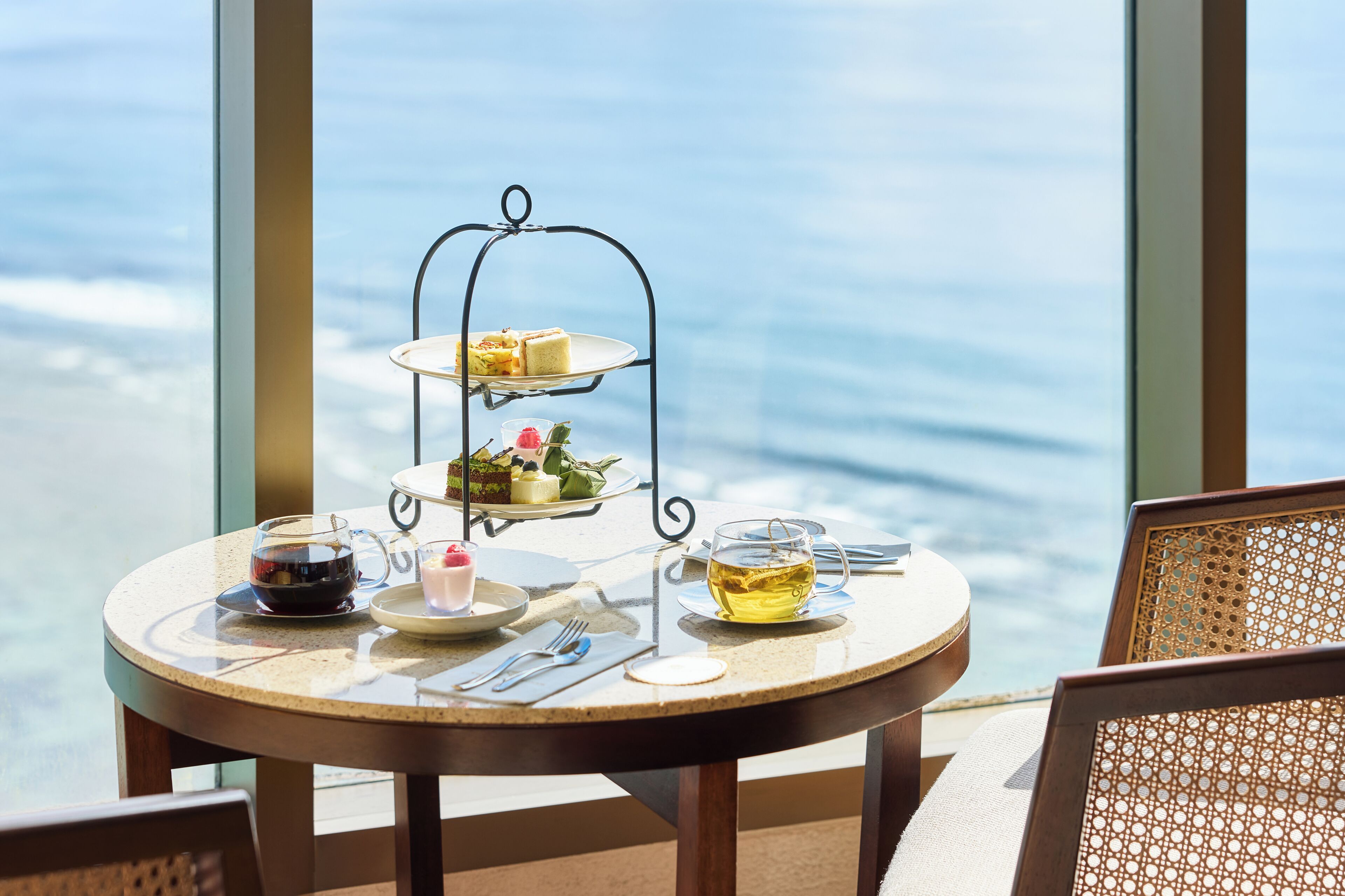 A round table with a tiered dessert stand, a glass teapot, and a cup of tea, set against a view of the sea.