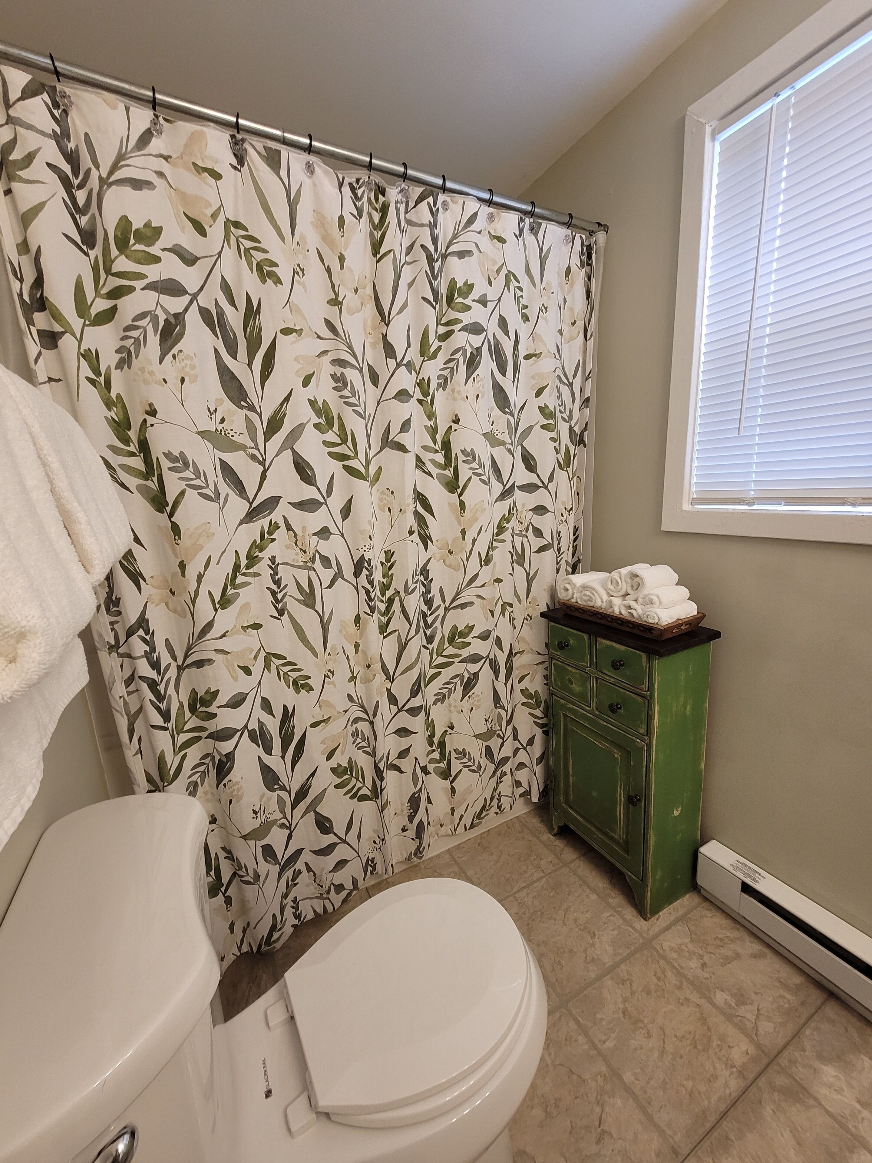 Casa de banho com cortina de chuveiro estampada com flores, sanita, armário verde com toalhas e janela com persianas.