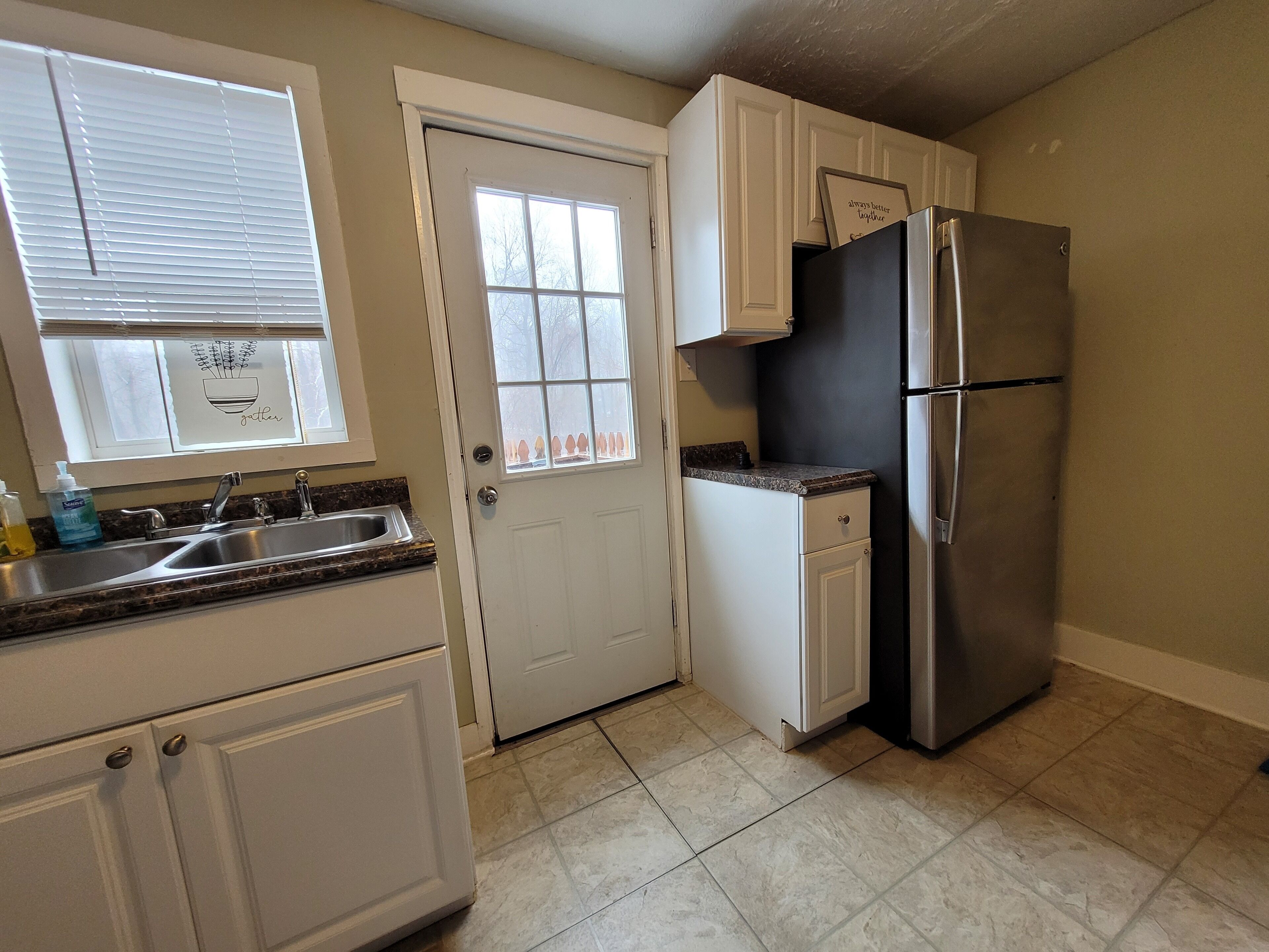 A kitchen with white cabinets, a stainless steel refrigerator, a window with blinds, and a sink with a faucet.