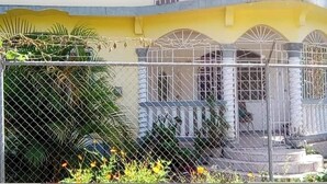 A two-story building with a yellow facade, arched windows, and a fenced courtyard. - Agatha's Home In Paradise  (Westmoreland Parish)