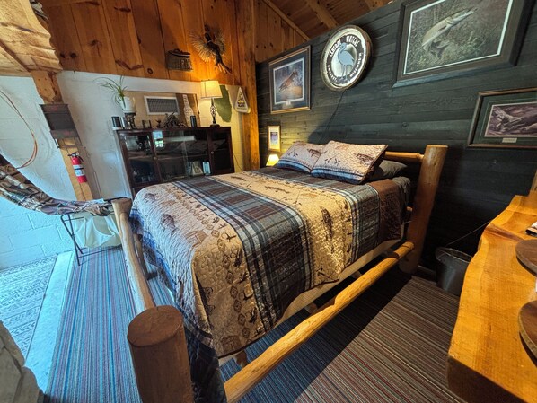 Una habitación acogedora con una cama de madera, un escritorio pequeño y una silla. Hay cuadros en la pared y una lámpara sobre el escritorio.