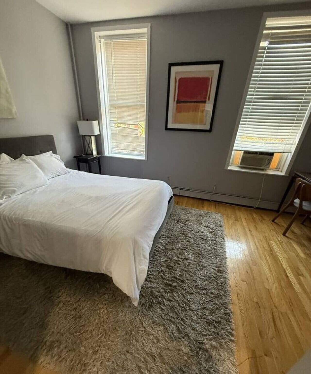 A bedroom with a bed, a gray rug, a nightstand, a lamp, a framed picture, and a window with blinds.