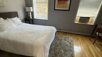 A bedroom with a bed, a gray rug, a nightstand, a lamp, a framed picture, and a window with blinds.